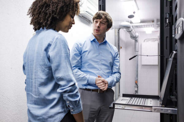 Ein Mann und eine Frau in einem Server-Raum im Keller vor einem aufgeklappten Laptop.