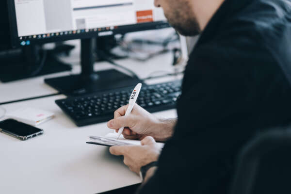 Eine Person am Schreibtisch in seitlicher Ansicht über die Schulter. Die Person schreibt etwas mit einem Kugelschreiber auf einen Block.